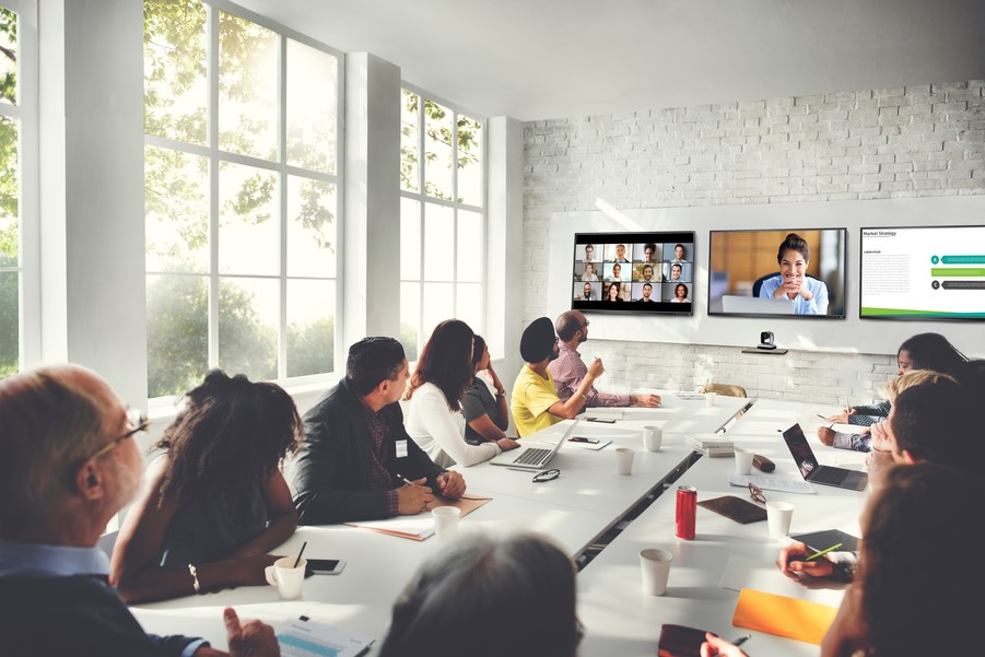 A video conference meeting around a large table in a windowed meeting room with three screens in the front of the room. 